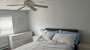 Ein Schlafzimmer mit einem Bett, einem Schminktisch, einem Nachttisch, einem Deckenventilator und einem Fenster mit Jalousien.