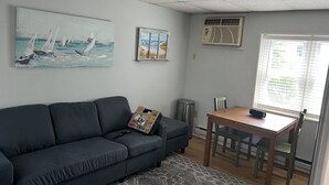 Ein Wohnzimmer mit einem dunkelgrauen Sofa, einem kleinen Holztisch mit Stühlen, einem Fenster mit Jalousien und zwei Wand-Klimaanlagen.