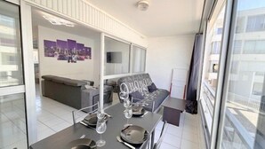 A modern living room with a sofa, a dining table set with plates and glasses, and a large window offering a view of the city.