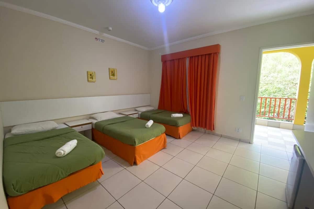 Habitación de hotel con tres camas, cada una con ropa de cama verde y reposapiés naranja. Hay una mesita y una silla, una ventana con cortinas naranjas y un balcón con barandilla roja.