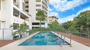 A swimming pool with a wooden deck, surrounded by a metal fence, and adjacent to a multi-story building with balconies. - ZEN AT ARKABA - 3BR Luxury Apartment (Darwin)