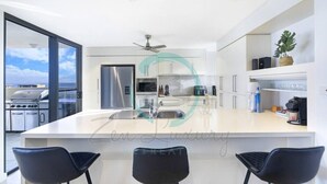 A modern kitchen with a large island, bar stools, and a view of the outdoors. - ZEN AT ARKABA - 3BR Luxury Apartment (Darwin)