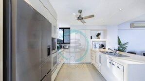 A modern kitchen with stainless steel appliances, a central island with a sink, and a ceiling fan. - ZEN AT ARKABA - 3BR Luxury Apartment (Darwin)