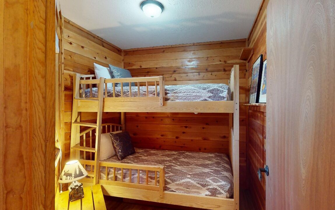 Quarto com beliche, paredes de madeira e uma pequena mesa de cabeceira com abajur.
