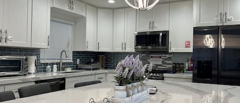 A modern kitchen with white cabinets, a central island, a stainless steel refrigerator, and a unique pendant light.