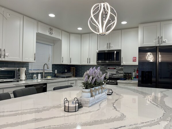 A modern kitchen with white cabinets, a central island, a stainless steel refrigerator, and a unique pendant light.