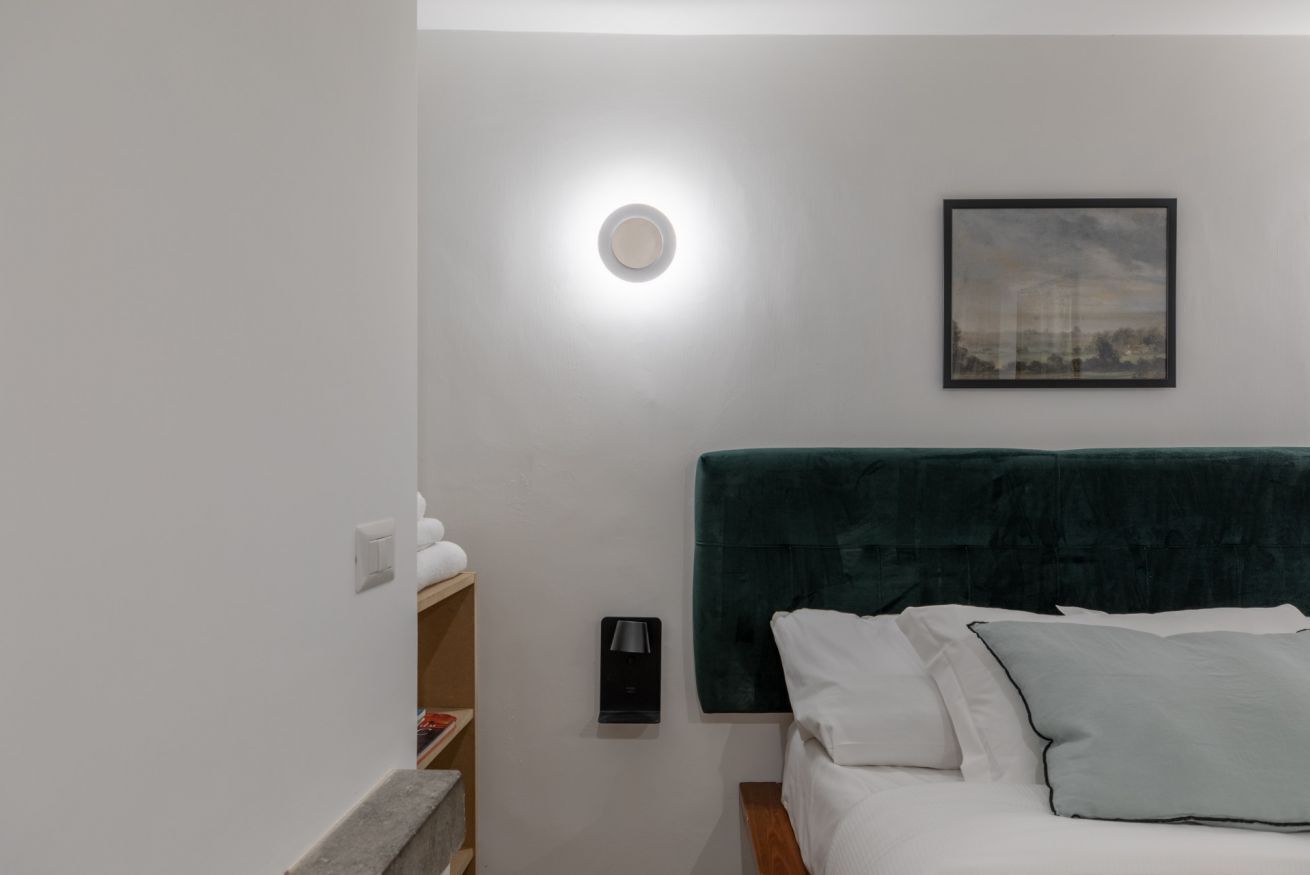 Quarto com cama, mesa de cabeceira, luminária na parede, quadro emoldurado e estante com livros.