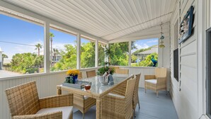 Un porche cubierto con mobiliario de mimbre, una mesa de vidrio y vistas a palmeras y casas.