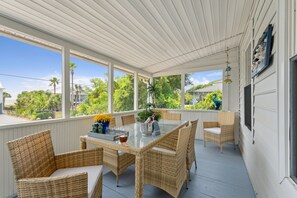 Eine überdachte Veranda mit Korbmöbeln, einem Glastisch und Blick auf Palmen und Häuser.