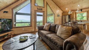 A wooden cabin interior with a large window, a brown leather sofa, a dining table, and a kitchen area. - Romantic retreat with spectacular mountain view. (Blue Ridge)