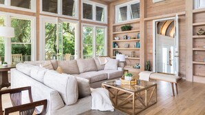 A spacious living room with a large beige sectional sofa, a glass coffee table, and wooden flooring. There are multiple windows providing natural light and a view of greenery outside.