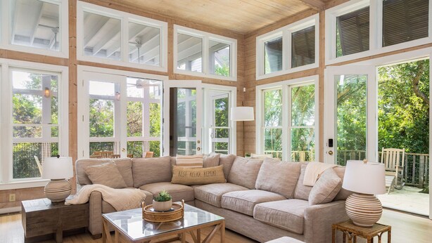 A spacious living room with a sectional sofa, a glass coffee table, and a wooden floor. Large windows allow natural light to fill the room.
