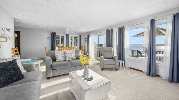 A living room with a sofa, armchair, and coffee table, a dining area with a table and chairs, and large windows with curtains. - Cape Cod Waterfront with Stunning Views (Buzzards Bay)