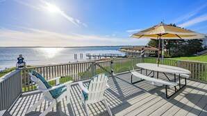 A deck with Adirondack chairs, a table, and an umbrella overlooking a body of water. - Cape Cod Waterfront with Stunning Views (Buzzards Bay)
