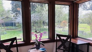 Une pièce d’angle dotée de trois grandes fenêtres, d’une chaise en bois, d’une petite table avec une plante et d’un luminaire suspendu.