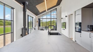 Een ruime kamer met grote ramen, een open haard, een keukenhoek en een zithoek met uitzicht op het omliggende landschap.