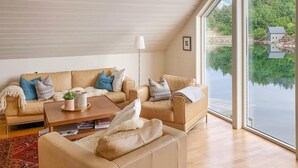 Uma sala de estar moderna com um sofá bege, uma mesa de centro e uma grande janela com vista para um lago e uma casa pequena.
