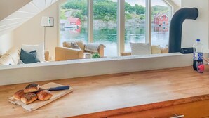 Quarto aconchegante com uma grande janela que dá para um corpo de água e uma casa vermelha. Há uma mesa de madeira com pão e uma faca, e uma garrafa de água no parapeito da janela.