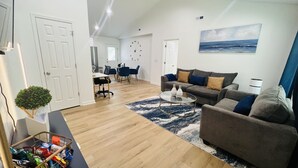 Un salon moderne avec un canapé gris, une table basse et un tapis. On aperçoit à l’arrière-plan un coin repas avec une table et des chaises.