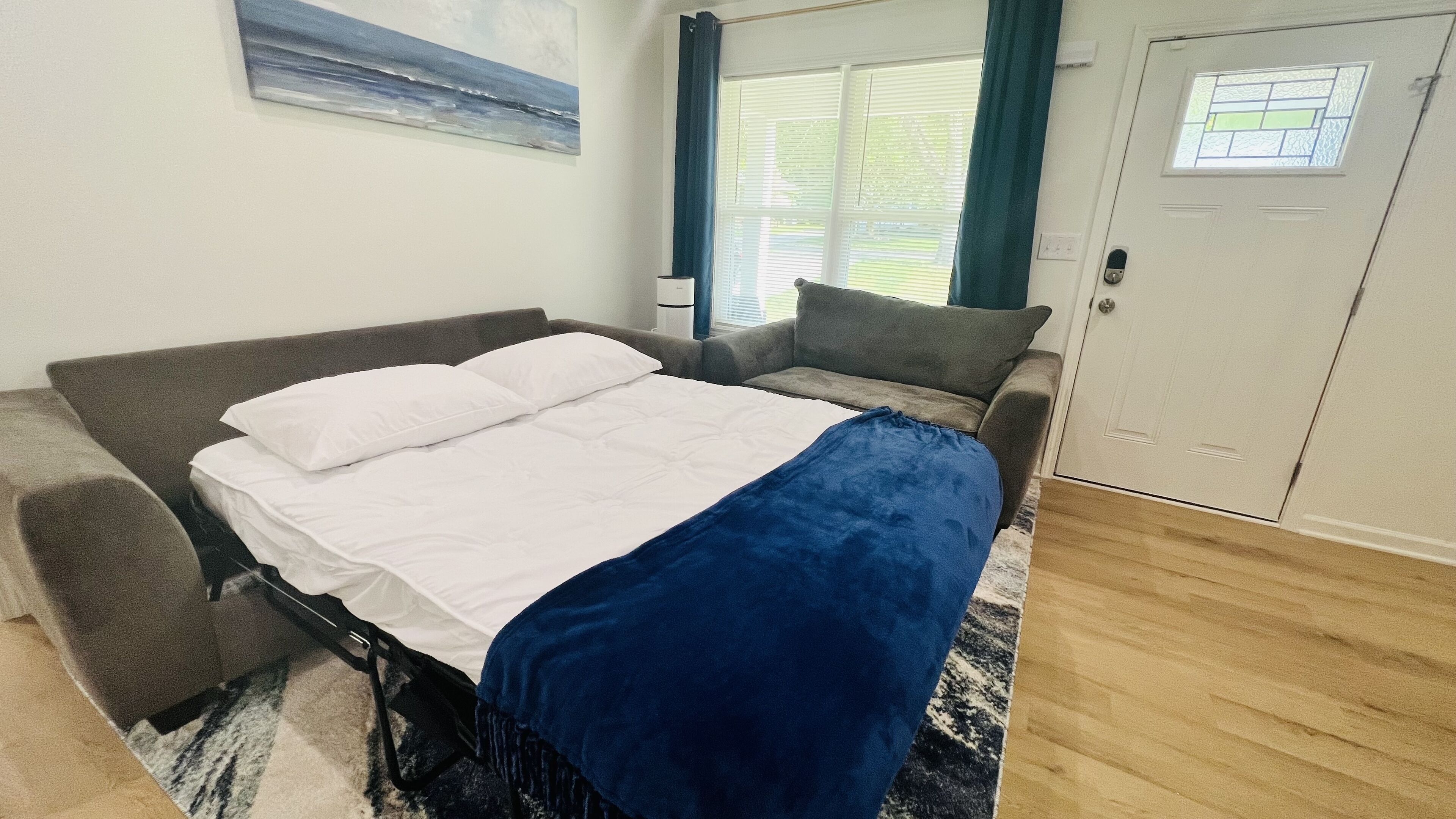 Un dormitorio con cama, sofá, ventana con cortinas, puerta y un cuadro en la pared.