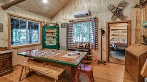 A cozy cabin interior with a wooden dining table, a green cabinet with animal figurines, a bed with a checkered curtain, and a window with a view of trees. - Quintessential Maine cabin w/ private waterfront (Augusta)