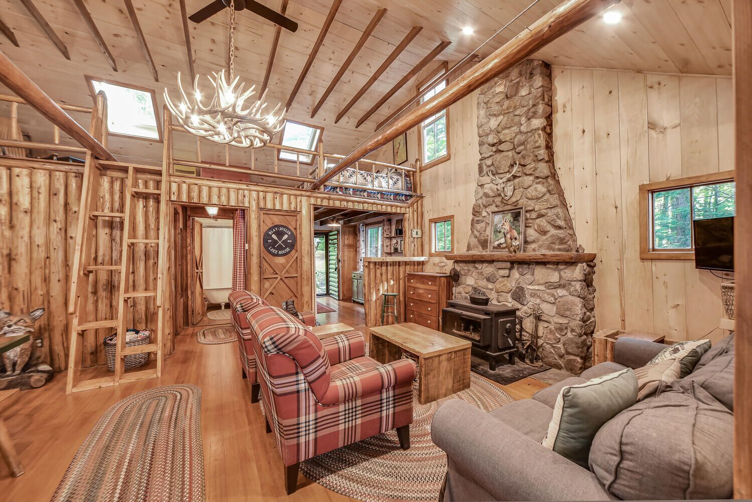 A rustic cabin interior with a stone fireplace, plaid sofas, and a wooden ceiling.