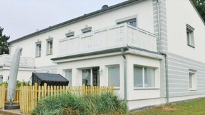 A two-story white building with a balcony, a wooden fence, and a grassy area. - Nilles Huuske - Luxusferienwohnung (Sellin)
