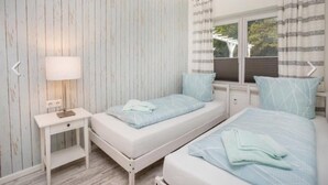 A bedroom with two beds, a nightstand, a lamp, and a window with curtains. - Nilles Huuske - Luxusferienwohnung (Sellin)