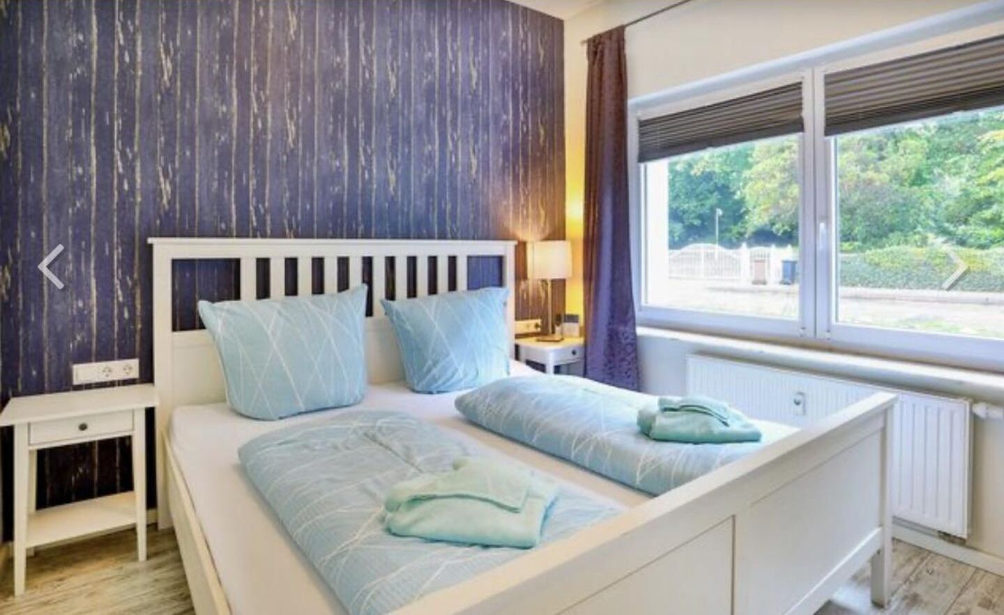 A bedroom with a white bed, light blue bedding, a nightstand, a lamp, and a window with a view of greenery.