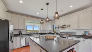 A modern kitchen with white cabinets, stainless steel appliances, a large island with stools, pendant lighting, and a view of the outdoors through a window.