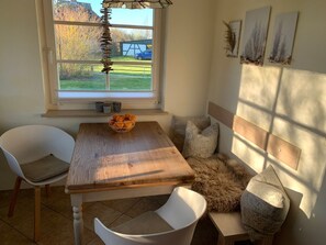A cozy dining area with a wooden table, white chairs, a bowl of oranges, and a bench with patterned cushions.