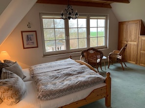 A cozy bedroom with a large bed, a wooden wardrobe, a wicker chair, and a view of the outdoors through a window.