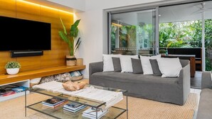 Ein modernes Wohnzimmer mit grauem Sofa, einem Glastisch und einem an einer Holzplatte befestigten Flachbildfernseher. Ein großes Fenster bietet Blick auf grüne Vegetation.