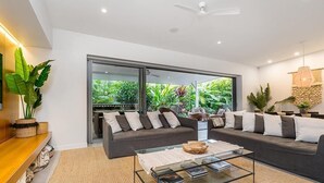 Ein modernes Wohnzimmer mit einem Glastisch, zwei Sofas mit Kissen und einem Deckenventilator, mit Blick auf Grünflächen.