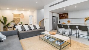 Ein modernes Wohnzimmer mit grauem Sofa, einem Glastisch und einer Küchenzeile mit Edelstahlkühlschrank und Barhocker.