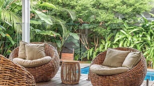 Korbstühle mit Kissen auf einer Holzterrasse neben einem Pool, umgeben von üppiger Bepflanzung.