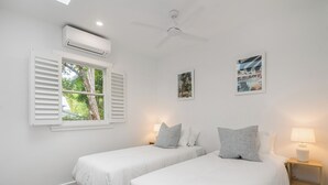 Une chambre avec deux lits, un ventilateur de plafond, un appareil de climatisation, une fenêtre avec des volets et une table de chevet avec une lampe.