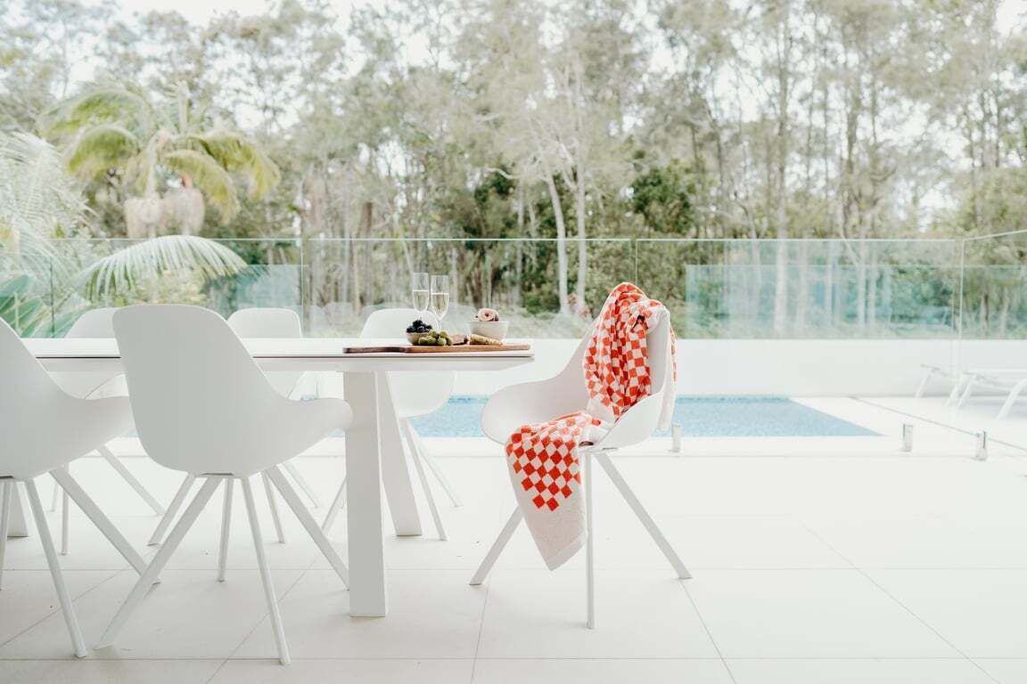 A modern outdoor dining area with white chairs and a table, overlooking a swimming pool and lush greenery.