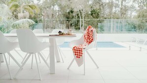 Un espace repas extérieur moderne, avec des chaises blanches et une table, offrant une vue sur une piscine et une végétation luxuriante.