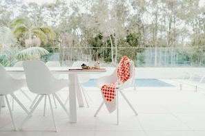 Uma área de refeições moderna ao ar livre, com cadeiras brancas e uma mesa, com vista para uma piscina e vegetação exuberante.