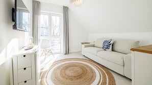 Ein stilvolles, helles Zimmer mit einem weißen Sofa, einem an der Wand montierten Fernseher, einem Schreibtisch mit Lampe und einem großen Fenster mit Vorhängen.