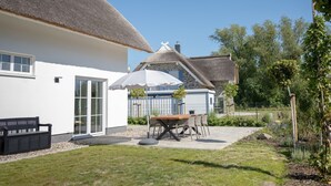 Ein Haus mit einem Strohdach, einer Terrasse mit Tisch und Stühlen sowie ein Garten mit einer Bank.