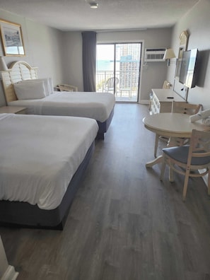A hotel room with two beds, a dining table, chairs, a TV, and a balcony with a view of buildings.