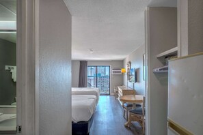A hotel room with a bed, a desk, a chair, a window with a view of buildings, and an air conditioning unit.