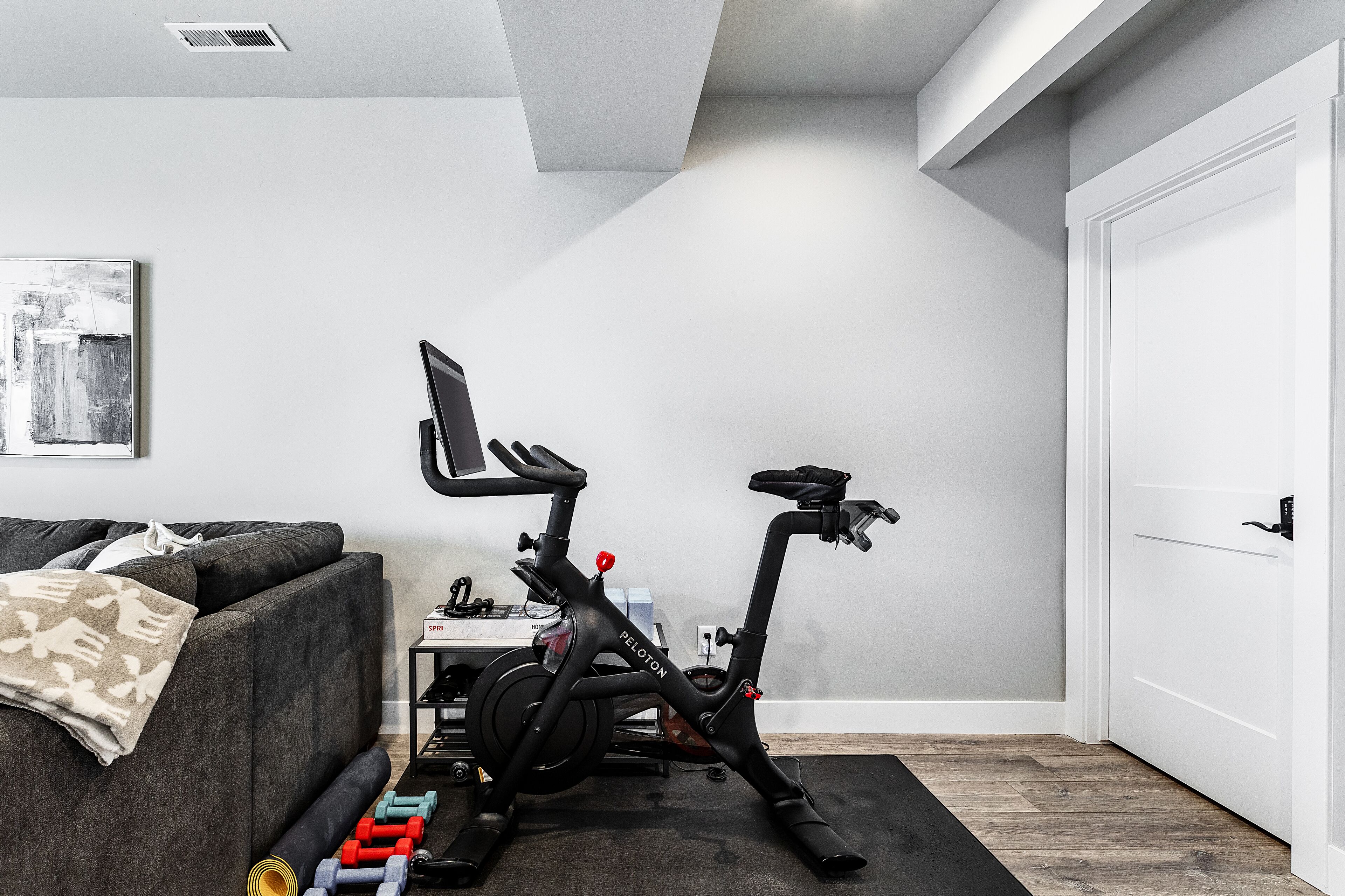 A modern living room with a grey sofa, a standing exercise bike, and a small table with dumbbells.