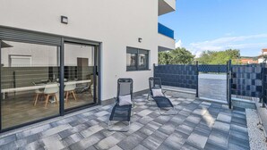 Une terrasse extérieure moderne, dotée d’une porte vitrée donnant sur un espace cuisine, de deux fauteuils et d’un sol carrelé.
