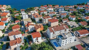 Vue aérienne d’un quartier résidentiel situé sur la côte, avec des bâtiments dotés de toits recouverts de tuiles rouges, une piscine et une vue dégagée sur la mer.