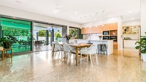 A modern kitchen with a central island, a dining area with white chairs, and a large sliding glass door leading to an outdoor area. - ZENLUX - 5BR Holiday Home with Pool (Fannie Bay)