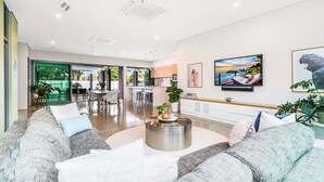 A spacious living area with a large sofa set, a coffee table, and a television mounted on the wall. There is a dining area visible through an open doorway. - ZENLUX - 5BR Holiday Home with Pool (Fannie Bay)