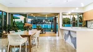 A modern kitchen with a marble countertop, a dining area with wicker chairs, and a view of a garden through large windows. - ZENLUX - 5BR Holiday Home with Pool (Fannie Bay)
